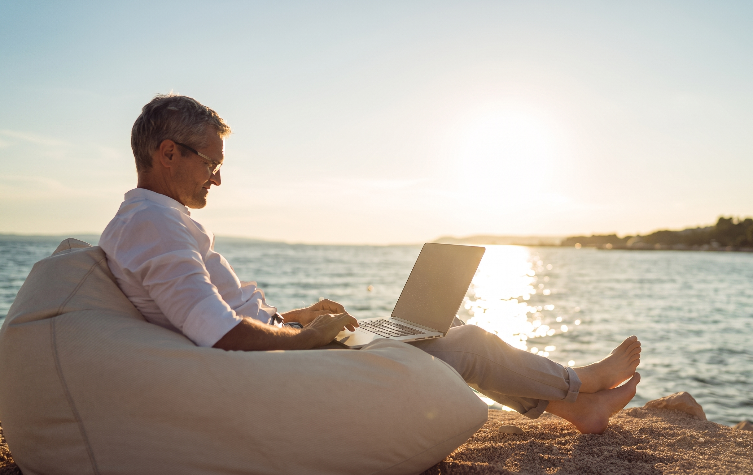 Middle-aged man working on a laptop while relaxing in a bean bag chair on a sunny beach at sunset.