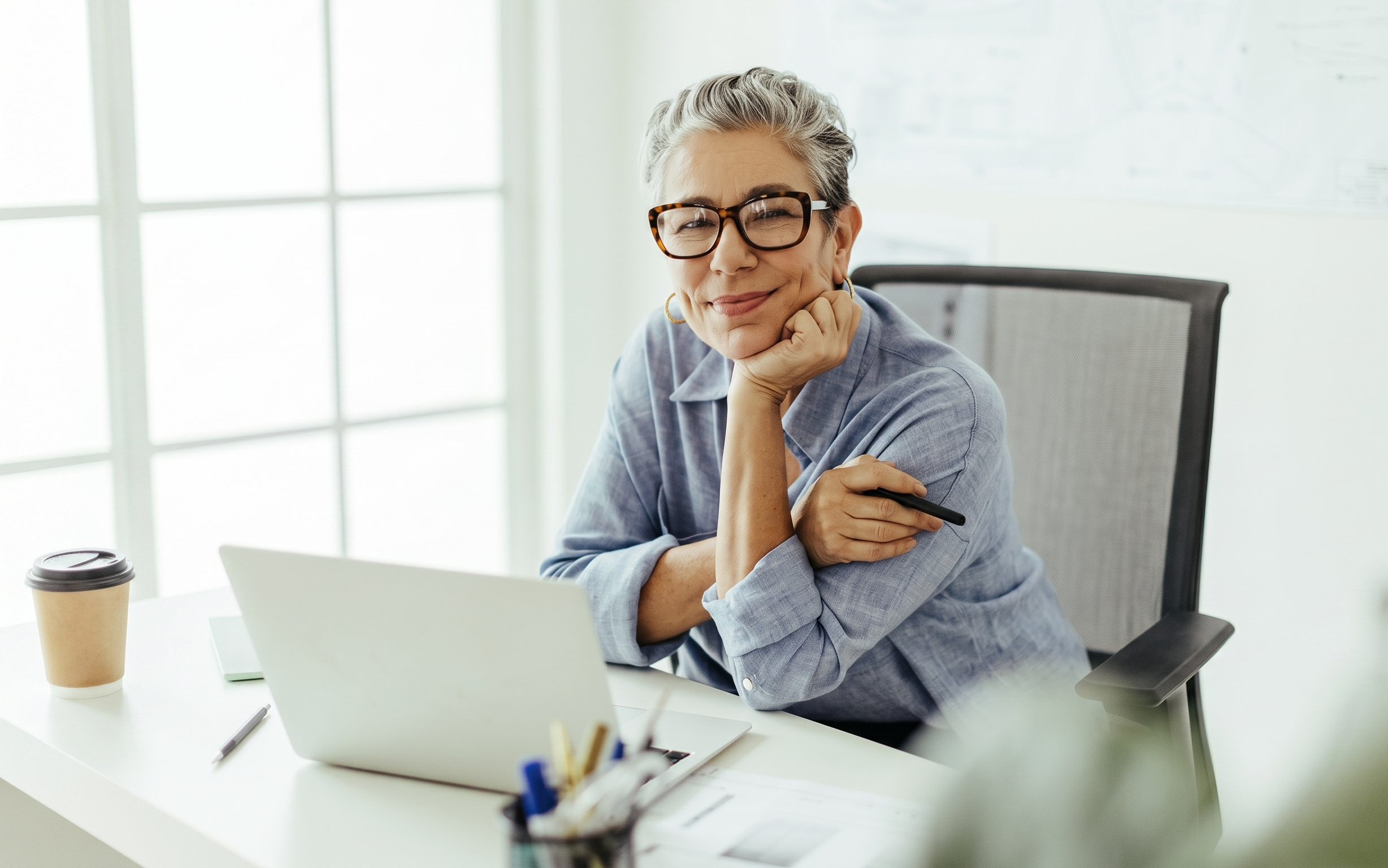 An elegant mature woman with grey hair and glasses smiling at her office desk with a laptop